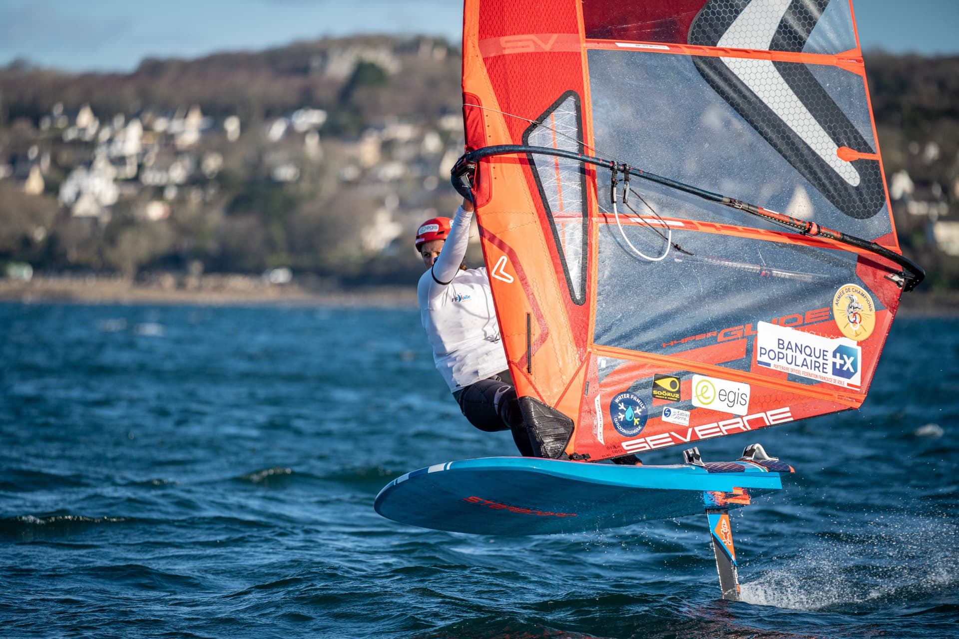 A windsurfer on an iQFOiL foiling board with an orange sail rides above the water off Brest, France, February 2023.